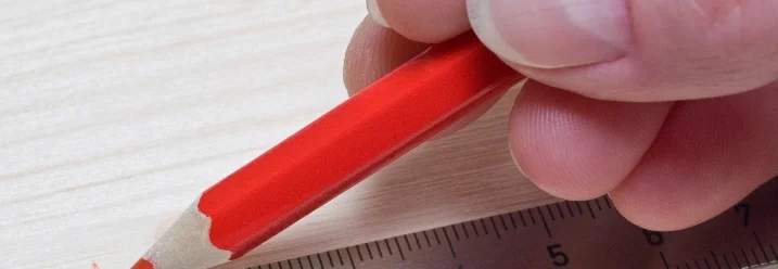 Eine Hand zeichnet mit einem roten Stift eine Linie an einem Metalllineal entlang.